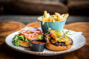 Cheese burger and bun with lettuce, tomato and onion on a round plate with side dish of fries and pot of tomato sauce on a wooden table top, served in a pub or restaurant, with shallow depth of field